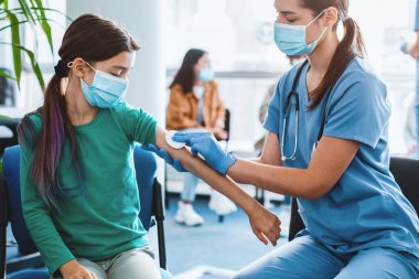 Küresel salgın sırasında yeni modern grip aşısı için klinik deney. Tıbbi maske takan genç kız hastanede iğne oluyor. Doktor hastayı iğneye hazırlıyor. Kolunu pamukla ovuyor.