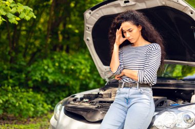 Şaşkın genç kadın sürücü açık araba kaputuna yaslanıyor, yolun ortasında araba çarpıyor. Gergin Ortadoğulu bayan araba servisi bekliyor, yakın plan fotoğraf, kopyalama alanı