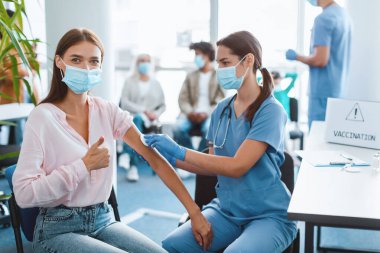 Koruyucu yüz maskesi takan genç bir kadın hastanede iğne yaptırıyor. Modern bir klinikte koluna pamuk pedi takmadan önce deriyi dezenfekte eden doktor.