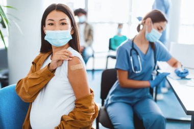 Grip aşısı. Aşılanmış Asyalı Kadın Coronavirus antiviral enjeksiyonundan sonra koluna bant takmış klinikte koluna sarılmış bir şekilde kameraya gülümsüyor. Grip Virüsü Koruma