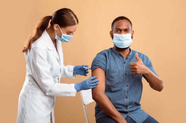 Koruyucu maskeli Afro-Amerikalı adam grip aşısı sırasında baş parmağını kaldırarak bej rengi stüdyo geçmişine yapılan bağışıklığı onaylıyor. Genç doktor erkek hastaya aşı yapıyor.