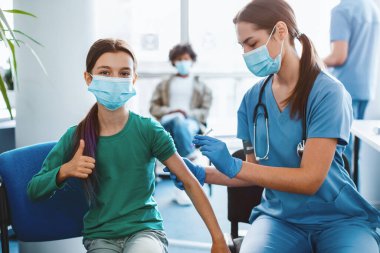 Başarılı grip aşısı konsepti. Omzuna aşı enjekte edilirken başparmağını hareket ettiren, klinik hastanesinde poz veren, geçmişi bulanıklaştıran olumlu maskeli kız. Dokunulmazlık