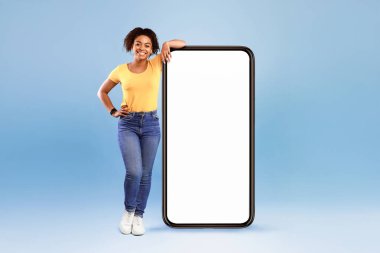 Büyük beyaz ekranlı akıllı telefona yaslanan mutlu Afro-Amerikalı kadın, mavi arka plandaki kameraya gülümsüyor, mobil uygulama, web sitesi, reklam tasarımı