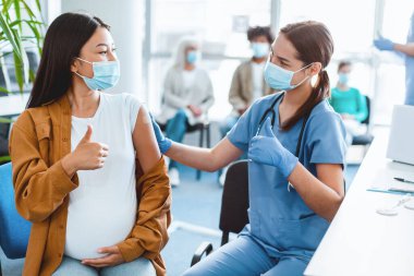 Aşı akışı 19. Aşılanmış Asyalı Kadın ve Kadın Doktor Klinikte koruyucu maske takarak birbirlerine bakarken başparmaklarını havaya kaldırıyorlar. Hemşire ve Hasta Bağışıklığı Onaylıyor