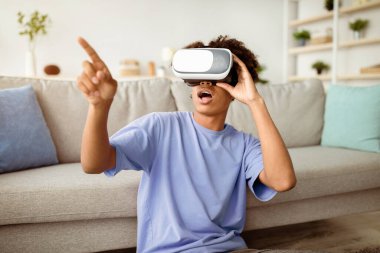 Çağdaş ev eğlenceleri. Sanal gerçeklik kulaklığı takmış Afro-Amerikalı genç adam içeride siber uzayı keşfediyor. VR gözlüklü siyah ergen etkileşimli bilgisayar oyunu oynuyor