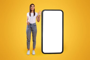 Cool Mobil Teklif. Büyük, beyaz ekranlı, kocaman bir cep telefonunun yanında duran mutlu sıradan bir kadın, tamam gösteriyor, cep telefonu için harika bir uygulama ya da web sitesi öneriyor, turuncu duvar, model al, tüm vücut uzunluğu