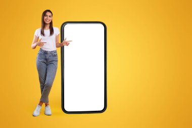 Mobil Uygulama Reklamı. Portakal Stüdyosu 'nun arka planında duran kocaman beyaz akıllı telefon ekranını gösteren mutlu kadının tüm vücut uzunluğu. Şuna bak, cep telefonu görüntüsü.