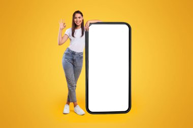 Büyük beyaz ekranlı, büyük akıllı telefona yaslanan ve işaretli neşeli bayan yeni uygulama ya da web sitesi öneriyor, sarı arka planda duruyor, görüntüyü taklit ediyor, vücut uzunluğunu arttırıyor.