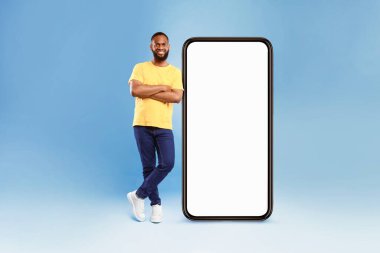 Mobil uygulama, web sitesi veya çevrimiçi reklam tasarımı için modelleme yapan, mavi stüdyo arka planında büyük boş akıllı telefona yaslanan, tam boy Afro-Amerikalı genç adam.