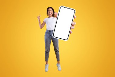 Cool Mobil Teklif Reklamı. Portakal rengi stüdyo arka planda havaya sıçrayan büyük boş bir cep telefonu ekranı tutan neşeli genç bayan, barış işareti hareketi gösteriyor, bedava fotokopi alanı, dikey