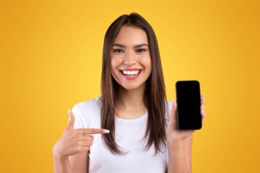 Bu havalı uygulamayı kullan. Siyah boş ekranlı akıllı telefonu tutan neşeli genç bayan, mobil uygulama reklamı için boş ekrana işaret ediyor, cihaz modeli, turuncu stüdyo arka planı