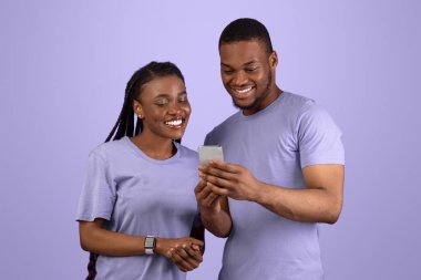 Mutlu siyah çift cep telefonu ekranına bakıyor, yeni bir uygulama kullanıyor, menekşe rengi mor stüdyo arka planı. Neşeli Afrikalı Amerikalı adam, modern eğlence anlayışı olan bayanla akıllı telefonu paylaşıyor.