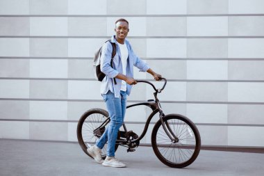 Sırt çantası ve modern bisikleti olan yakışıklı bir zenci. Şehir sokağındaki tuğla duvarın yanında. Pozitif Afrikalı Amerikalı bisikletçi şehir kültürünü keşfediyor, spor yapıyor. Sağlıklı yaşam tarzı