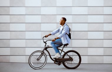 Sırt çantalı ve kulaklıklı heyecanlı siyah adam dışarıda tuğla duvarın yanında bisiklet sürüyor.