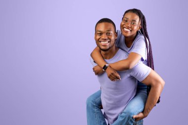 Sırtında taşı. Gülümseyen Afrikalı Amerikalı bir adamın portresi sırtında taşıyarak ve sırtında taşıyarak, birlikte zaman geçirerek, mor stüdyo arka planında tek başına dikilip, eğlenerek