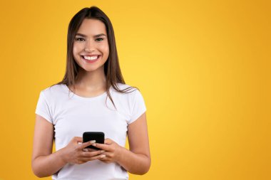 Halk ve Teknoloji Konsepti. Portakal rengi stüdyo arka planında izole edilmiş akıllı telefon kullanan sıradan gülümseyen genç kadın portresi kameraya bakıyor. İnternette sohbet eden kadın, sosyal medyada gezinen.