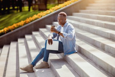 Tabletli mutlu siyah adam şehir merkezindeki taş merdivenlerde kahve içiyor.