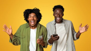 Mobil Teklif. Bej rengi stüdyonun arka planında poz verirken sevinçten havalara uçan, internetteki reklamlara duygusal tepki veren iki heyecanlı siyahi arkadaşın portresi.