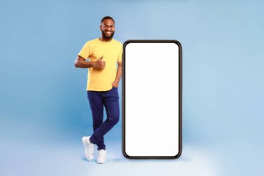 Güzel mobil uygulama. Mavi arka planda dev akıllı telefonun yanında el kol hareketi yapan neşeli Afrikalı Amerikalı adam web sitesi için model ya da boş ekranda yeni bir uygulama.