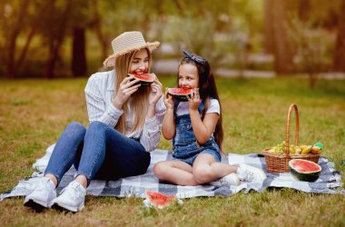Anne ve küçük kızı güzel yeşil parkta battaniyenin üzerinde oturmuş karpuz yiyen aile pikniğinin tadını çıkarıyorlar. Ana Konseptle Hafta Sonu