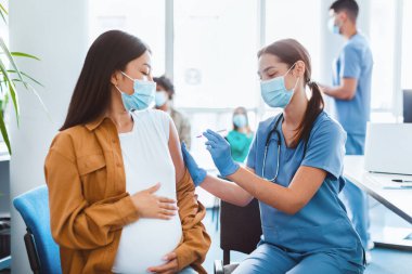 Kadın doktor elinde şırınga tutarak hamile kadının omzuna 19 aşıyı enjekte ediyor. Asyalı kadın sarılma göbeği. Grip aşısı klinik deneyleri, korona virüsü tedavisi yan etkileri