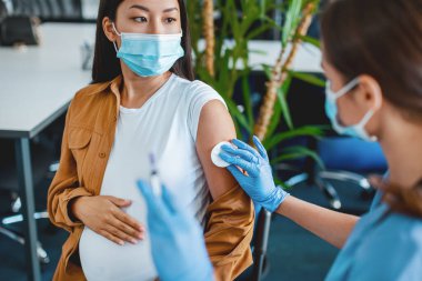 Koruyucu maskeli kadın doktor ve eldivenler hamile hastaya covid antiviral aşı enjekte etmeye hazırlanıyor, elini pamukla ovuyor. Kitle aşılama merkezi, küresel aşılama kampanyası