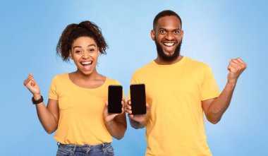 Harika bir mobil uygulama. Heyecanlı Afrikalı Amerikalı çift akıllı telefonları modelli gösteriyor, EVET işareti yapıyor, mavi stüdyo arka planı üzerinden yeni web sitesi reklamı yapıyorlar. Büyük kazanç, büyük satış, büyük şans konsepti.