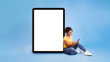 Cep telefonu kullanan Afrikalı genç bir bayan beyaz ekranlı, mavi arka planlı dev tabletin yanında oturuyor. Uygulama veya web sitesi için model, reklam tasarımı için alan