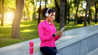 Kamu parkında dinlenirken akıllı telefon için spor uygulaması kullanan siyah kız koşucu, boş alan