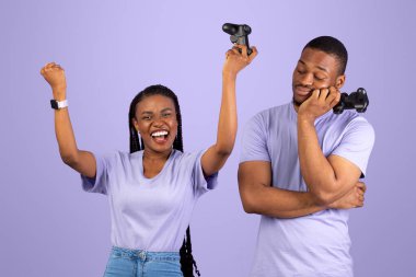 Modern online oyunda kazanma ve kaybetme duyguları. Heyecanlı Afrikalı Amerikalı kadın, elinde joystick ile yumruklarını sallayarak başarıya seviniyor. Morarmış arka planda izole edilmiş üzgün bir adam ona bakıyor.