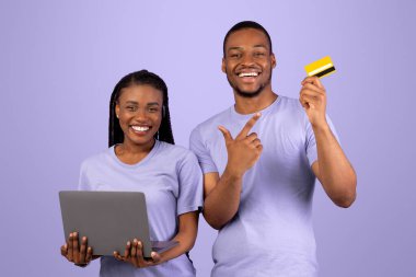 Çevrimiçi Kolay Bankacılık ve E-Ticaret. Mutlu siyah çift dizüstü bilgisayar kullanıyor, neşeli adam tutuyor ve Violet stüdyosunun arka planında izole edilmiş kredi kartını işaret ediyor, bilgisayarla online alışverişler yapıyor.