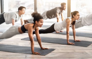 Stüdyoda yoga yapan bir grup genç sporcu, tahta falankasana pozunda duruyorlar, sağlıklı yaşam tarzının tadını çıkarıyorlar, yandan bakıyorlar.