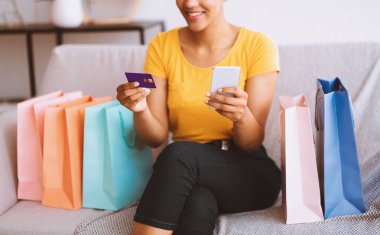 Çevrimiçi Alışveriş Konsepti. Elinde akıllı telefon ve kredi kartı olan tanınmamış Afrikalı kız koltukta çantalarla oturuyor.