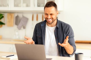 Sanal iş toplantısı. Çevrimiçi iş arkadaşlarıyla sohbet eden neşeli genç adam, dizüstü bilgisayarlı webcam ile konuşan, evdeki mutfağın içinde oturan, boş alan