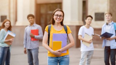 Dışarıda üniversite binasının yanında duran, sırt çantası ve kitaplarıyla poz veren, kameraya gülümseyen öğrenci kız.