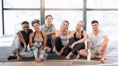 Yoga eğitiminden sonra spor salonunda poz veren mutlu insanlar minderlere oturup gülümsüyor ve sağlıklı yaşam tarzının tadını çıkarıyorlar.