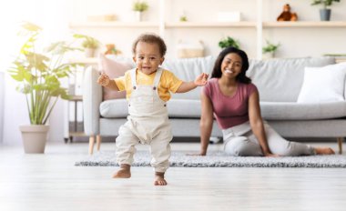 İlk Basamaklar. Sevimli bebek oturma odasında yürüyor, tatlı Afrikalı Amerikalı çocuk yere basıyor ve kameraya bakıyor, gururlu annesi arka planda gülümsüyor,