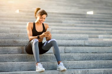 Sportif siyah kız dinlenirken su ve telefon kullanıyor, parkta eğitim görüyor, fotokopi çekiyor, güneş parlıyor.