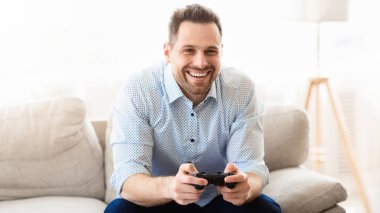 Genç adam evde video oyunu oynuyor. Koltuğunda elinde joystickle oturan mutlu adam.
