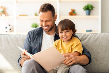 Hikaye zamanı. Genç, sevgi dolu bir baba, küçük oğluna masal okuyor, büyük çocuk kitabıyla evde oturuyor ve gülümsüyor.