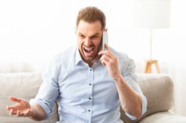 Sinirli adam cep telefonuyla konuşuyor, bağırıyor, kanepeye oturuyor, kopyalanmış.
