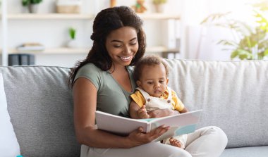 Anne şefkatinin portresi, evde koltukta dinlenirken sevimli küçük bir bebeğe kitap okuması, Afrikalı Amerikalı bir annenin bebeğiyle yakınlaşmasının yakın çekimi,