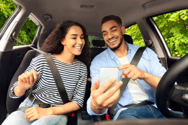 Neşeli orta doğulu sürücü güzel uzun saçlı kız arkadaşıyla güzel bir otomobille seyahat ediyor, cep telefonuna GPS koyuyor, akıllı telefon ekranına bakan çift, araba panelinden vurulmuş.