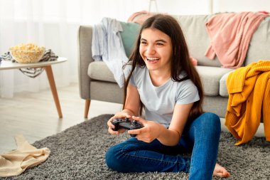 Covid tecrit sırasında dağınık bir odada oyun oynayan joystick 'li mutlu Hintli genç. Evde video oyunu oynayan kontrolörü olan neşeli bir ergen.