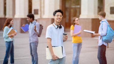 Dışarıdaki üniversite binasının yakınındaki çok ırklı öğrenciler arasında duran Asyalı öğrenci kameraya gülümsüyor. Daha Yüksek Öğrenim