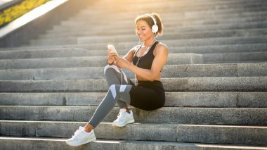 Güzel siyah kulaklıklı bayan koşucu parkta oturmuş, müzik dinliyor, telefon kullanıyor, mekanı kopyalıyor.