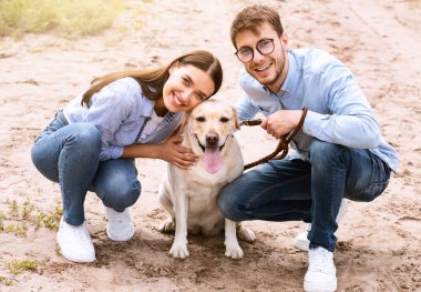 Köpek dostu randevu konsepti. Köpeğiyle oturan iki genç, kameraya bakıyor, tasmalı bir evcil hayvan tutuyor.