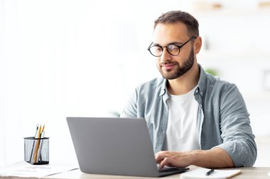 Gözlüklü genç beyaz adam internette çalışıyor, masa başında oturuyor ve ev ofisinden laptop kullanıyor. Millennial elemanı taşınabilir bilgisayarda, kapalı alanda uzak iş projesi yapıyor.