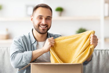 Karton paketleri açan mutlu beyaz adam başarılı alışverişlerden, yeni kazaklar çıkarmaktan, evden yeni şeyler almaktan memnun. Çevrimiçi mağaza ve dağıtım kavramı