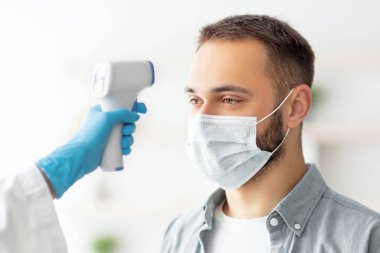 Lastik eldivenli doktor içeride kızılötesi termometreyle genç erkeklerin sıcaklığını ölçüyor. Coronavirus aşısı vurulmadan önce sağlık kontrolü yapan tıbbi maskeli bir adam. Çovid 'e karşı bağışıklık